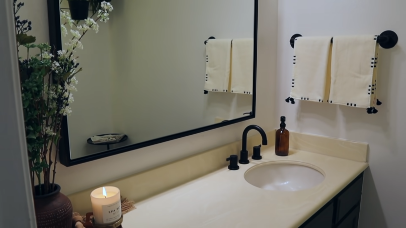 A Modern Bathroom Vanity Featuring a Large Rectangular Mirror, Matte Black Faucet Fixtures, Neatly Arranged Towels, a Lit Candle, and Decorative Plants